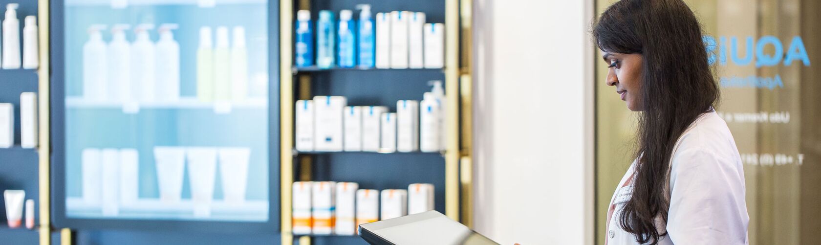 Eine Apothekerin im weißen Kittel arbeitet mit einem digitalen Tablet vor einem Regal voller medizinischer Produkte in der Offizin.