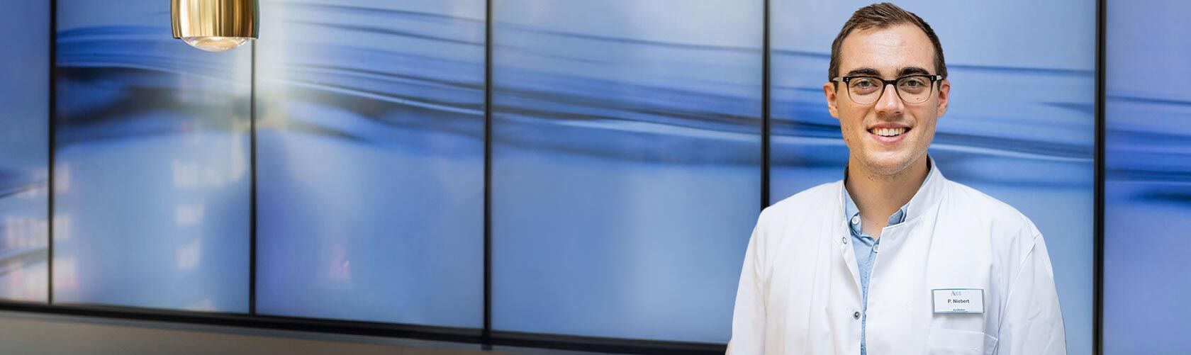 Ein lächelnder Mann im weißen Kittel steht vor einem modernen, blauen Hintergrund mit mehreren Bildschirmen.